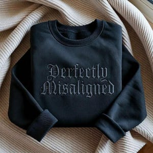 May include: Black sweatshirt with the embroidered text "Perfectly Misaligned" in a gothic font. The sweatshirt is folded, with the cuffs visible. The fabric appears soft and comfortable.