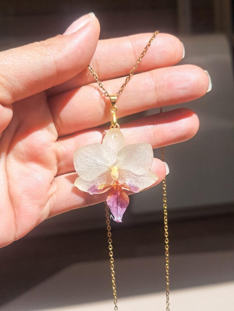 May include: A gold chain necklace with a delicate white and purple orchid pendant. The orchid is preserved in resin and has a natural, translucent appearance.