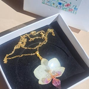May include: A gold chain necklace with a white and purple orchid pendant. The pendant is made of resin and features a real pressed flower. The necklace is in a white box with the text "Coco Floral Handmade Designs" on the lid.
