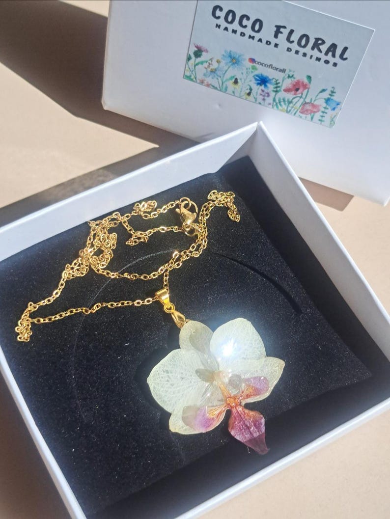 May include: A gold chain necklace with a white and purple orchid pendant. The pendant is made of resin and features a real pressed flower. The necklace is in a white box with the text "Coco Floral Handmade Designs" on the lid.