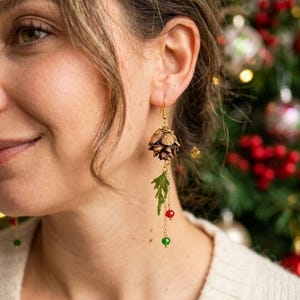 May include: A dangle earring featuring a small brown pinecone attached to a gold-tone hook. Below the pinecone hangs a green leaf-shaped charm, a small red bead, and a small green bead, all connected by a thin gold-tone chain.