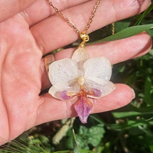 May include: A gold chain necklace with a white orchid pendant. The orchid has a purple center and is preserved in resin.