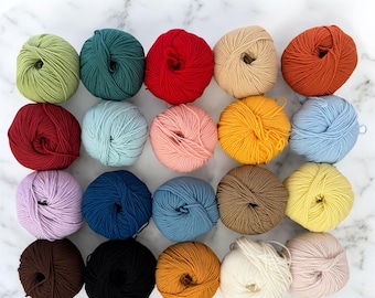 Könnte beinhalten: Zwanzig Wollknäuel in verschiedenen Farben, darunter Rot, Blau, Grün, Gelb, Braun und Weiß. Die Knäuel sind in einem Kreis auf einer weißen Marmorfläche angeordnet.