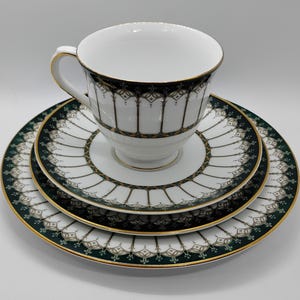 May include: A white teacup with a gold handle and rim sits atop a matching saucer and plate set. The set features a dark green and gold decorative pattern around the edges and on the cup.