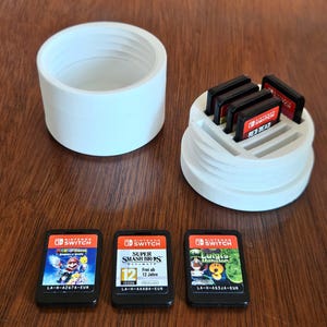 Op de afbeelding: Een witte 3D-geprinte Nintendo Switch game cartridgehouder met een bijpassend deksel. De houder bevat verschillende game cartridges, waaronder "Red Dead". Drie extra game cartridges worden weergegeven, waaronder "Super Smash Bros Ultimate".