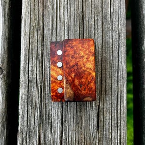 May include: A handcrafted wooden domino set with a rich, swirling grain pattern in shades of brown and orange. The dominoes are held together with silver-coloured magnetic closures. The set is displayed on a weathered wooden surface.