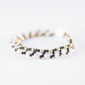 May include: A delicate bracelet made with black, gold, and clear beads. The bracelet is woven in a pattern that resembles a wave.