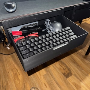 May include: A black keyboard, mouse, and red-framed glasses are in an open desk drawer. A stapler and other office supplies are also visible. The drawer is part of a black desk with a black mousepad and a Logitech mouse.