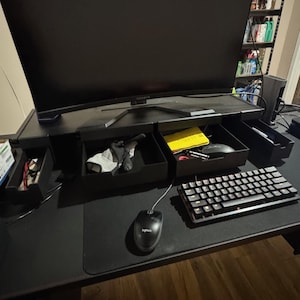 May include: A black computer desk with a large monitor, keyboard, and mouse. The desk has built-in drawers containing various items. A black and red mechanical keyboard sits to the right of the mouse.