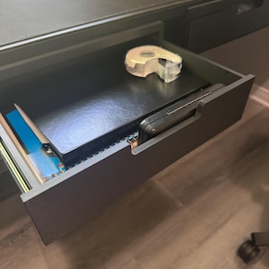 May include: A black desk drawer pulled open, containing a black notebook, a blue spiral-bound notebook, a black rectangular object, and a clear plastic tape dispenser with a roll of transparent adhesive tape resting on top of the black notebook.