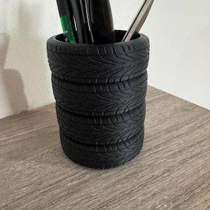 Może przedstawiać: Czarny organizer biurkowy w kształcie opony z zielonym długopisem, czarnym długopisem i srebrnym otwieraczem listów w środku.