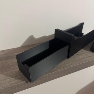 K&ouml;nnte beinhalten: Ein schwarzer, rechteckiger Organizer mit einem langen, offenen Fach und einem erh&ouml;hten R&uuml;ckenteil. Der Organizer besteht aus einem matten Material und steht auf einer hellen Oberfl&auml;che. Das Design ist minimalistisch und modern.
