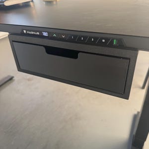 May include: A black adjustable standing desk with a smooth surface and a built-in drawer. The front edge features a digital control panel with the text MAGNUS, height display, arrow buttons, three memory presets, an S button, and a power indicator.