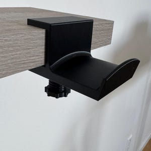 Puede incluir: Abrazadera de escritorio de metal negro con un gancho curvo para colgar objetos. La abrazadera está unida a una superficie de madera.