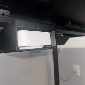 May include: A black under-desk mount holding a white device. The mount is rectangular and fixed to the underside of a black desk. The white device has a rounded top and a series of small holes.