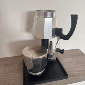 Puede incluir: Una cafetera de espresso de plata y negro con un mango negro y una base negra. La cafetera está sentada en una bandeja negra.