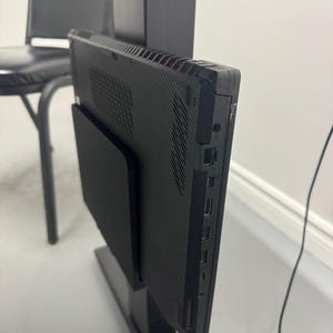 Könnte beinhalten: Ein schwarzer Computermonitor mit strukturierter Rückseite ist auf einem schwarzen Ständer montiert. Das Seitenprofil zeigt verschiedene Anschlüsse und Steckverbinder. Im Hintergrund ist ein schwarzer Stuhl zu sehen.
