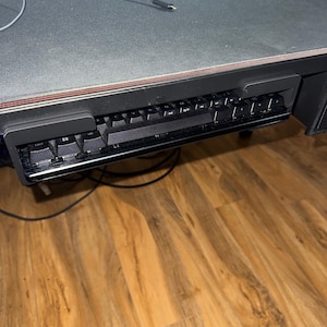 May include: A black mechanical keyboard is mounted under a desk. The keyboard has black keycaps and a black frame. The desk has a black surface with a red trim. The floor is made of wood.