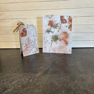 May include: A handmade greeting card and gift tag set. The card and tag feature a floral design with a butterfly accent. The color palette includes shades of orange, pink, and beige. The tag has a twine string for attaching to a gift.