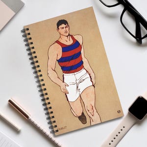 May include: A spiral-bound notebook with a kraft paper cover, depicting a man in a red, blue, and white athletic outfit. The notebook has a black spiral binding. A pen, glasses, and a smartwatch are also visible.