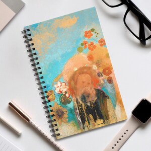 May include: Spiral-bound notebook featuring an artistic illustration of a bearded figure with a halo, surrounded by flowers. The cover has a blue and orange background with a gold accent. The notebook is open, displaying the spiral binding.
