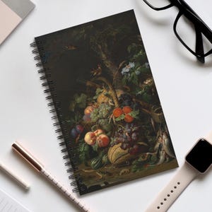 May include: Spiral-bound notebook with a still-life painting of a basket of fruit, grapes, and fish. The artwork has a dark background with green, red, and purple hues. The notebook is on a white surface, with a pen and watch visible.