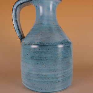Pode incluir: Um jarro de cerâmica com um esmalte azul claro e uma alça azul escura. O jarro tem um corpo arredondado e um bico para servir. A superfície tem uma aparência texturizada com pequenas manchas escuras.