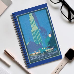 May include: A spiral-bound notebook with a blue cover featuring a vintage illustration of the Woolworth Building at night. The artwork is in shades of blue, green, and white, with the text "WOOLWORTH BUILDING JUNE NIGHT".