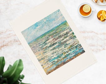 May include: A cream-colored kitchen towel featuring an abstract painting of the ocean. The artwork depicts the sea with blue, green, and white brushstrokes, suggesting waves under a blue sky. The towel is rectangular and lies on a marble surface.