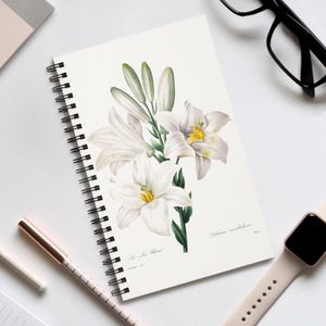 May include: A spiral-bound notebook with a botanical illustration of white lilies with yellow centres and green stems. The notebook has a black spiral binding and is on a white surface with a pen, watch, and glasses.