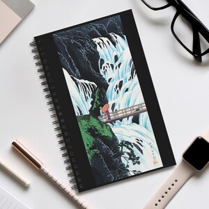 May include: Black spiral-bound notebook with a Japanese waterfall scene. The image shows a waterfall, a wooden bridge, and figures with an umbrella. The cover art is in shades of blue, green, and black.