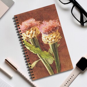 May include: Spiral-bound notebook with a watercolour illustration of two artichoke flowers. The flowers are yellow and pink, with green leaves against a brown background. The notebook is on a white surface.