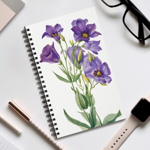 May include: A spiral-bound notebook with a watercolour painting of purple flowers with yellow centres and green leaves. A pen, glasses, and a smartwatch are visible. The notebook is open, displaying the floral artwork.