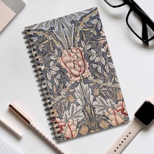 May include: A patterned spiral-bound notebook with a floral design in grey, pink, and cream. The cover displays stylized flowers and leaves. The notebook is open, showing the spiral binding and the patterned cover.