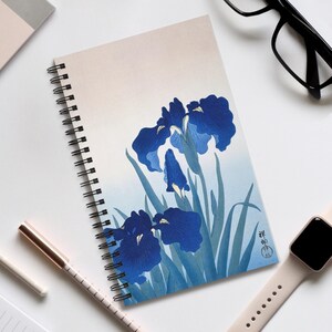 May include: A spiral-bound notebook with a blue iris floral design. The cover art shows a stylized illustration of irises with blue petals and green leaves. The notebook has a white background and a black spiral binding.