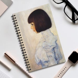 May include: Spiral-bound notebook with a portrait of a person with dark hair and a white ruffled collar. The artwork uses soft pastel colours. The notebook is on a white surface with a pen, watch, and glasses.