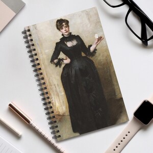 May include: Spiral-bound notebook with a portrait of a woman in a black dress. The artwork shows a woman holding a flower, set against a neutral backdrop. The notebook is on a white surface, with a pen, watch, and glasses.