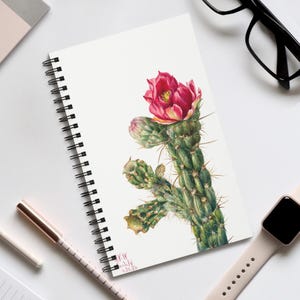 May include: A spiral-bound notebook with a watercolour illustration of a blooming cactus. The cactus is green with sharp spines and a vibrant red flower. The notebook has a white cover and a date written on the bottom.