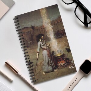 May include: Spiral-bound notebook featuring a classical painting of a woman in a long dress standing near a fire. The artwork depicts a mystical scene with a waterfall and a cauldron. The notebook has a black spiral binding.