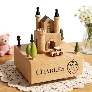 May include: A wooden music box featuring a castle design with two black-topped towers, a small car, and miniature trees. The box is engraved with the name "CHARLES" and a strawberry illustration. The music box sits on a white lace doily.