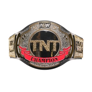 May include: A championship wrestling belt featuring a large circular center plate with the text TNT in gold lettering. A red banner below reads CHAMPION in black text. The silver plate includes intricate engravings and an AEW logo at the top.