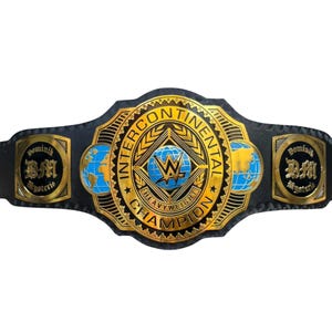 Puede incluir: Un cinturón de campeonato de lucha libre negro con una gran placa central dorada con el texto INTERCONTINENTAL HEAVYWEIGHT CHAMPION, un globo terráqueo azul y el logo de la WWE. Dos placas laterales muestran el texto Dominik Mysterio y las iniciales DM.