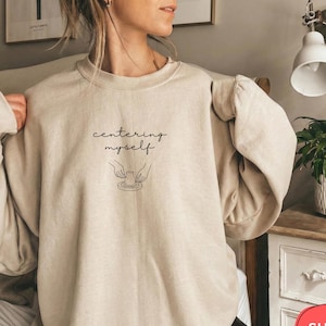 May include: Beige sweatshirt with the text "centering myself" above a line drawing of hands shaping pottery. The long-sleeved top has a crew neck and a relaxed fit. The image includes a red and white "SHIPS FAST!" sticker.