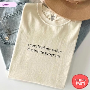 May include: Ivory-colored t-shirt with the text "i survived my wife's doctorate program" in black. The shirt is folded, with a hat and jeans in the background. The shirt has a Comfort Colors tag.