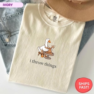 May include: An ivory short-sleeve t-shirt featuring a graphic of a white goose wearing an apron and working at a pottery wheel. Below the graphic, the text reads i throw things in a black serif font. The shirt has a Comfort Colors brand label.