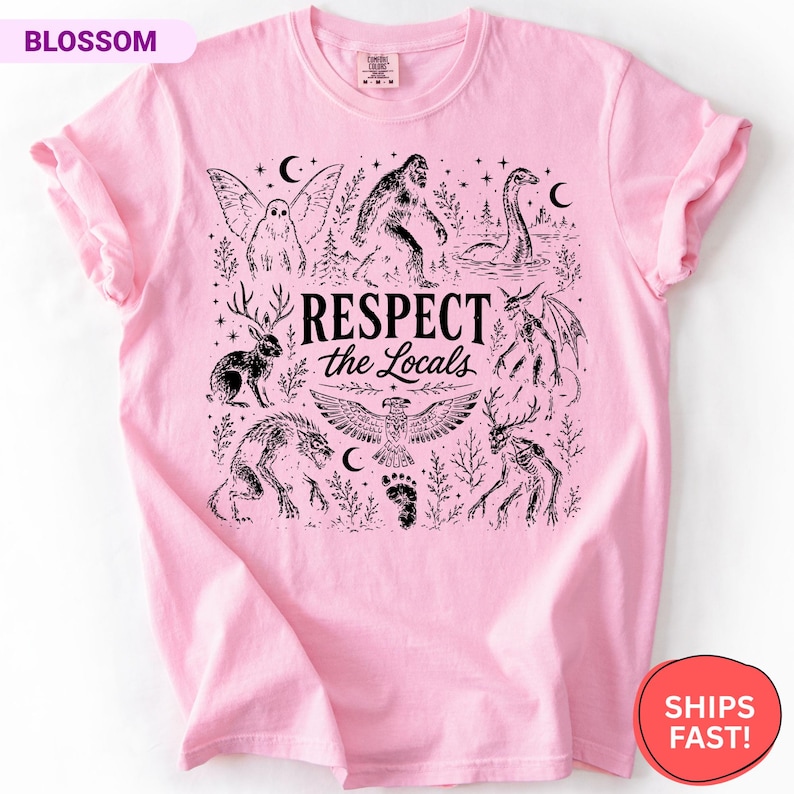 May include: A light pink t-shirt featuring a black graphic print of various mythical creatures including Bigfoot, a jackalope, and a mothman. The text "RESPECT the Locals" is centered in the middle, surrounded by stars, moons, and a footprint.