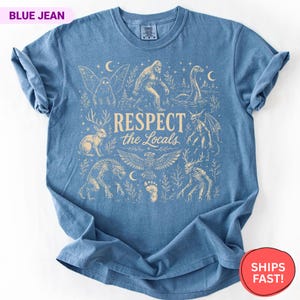 May include: A blue jean colored t-shirt featuring a cream-colored graphic print of various mythical creatures including Bigfoot, a jackalope, and a dragon. The central text reads "RESPECT the Locals" surrounded by stars, moons, and foliage.