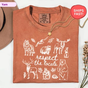 May include: Rust-colored t-shirt with a white graphic of various animals and the text "respect the locals." The shirt features a crew neck and a Comfort Colors tag. A gold star pendant necklace is draped over the shirt.