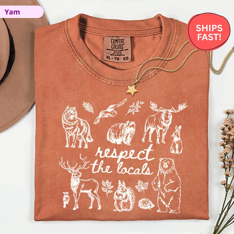 May include: Rust-colored t-shirt with a white graphic of various animals and the text "respect the locals." The shirt features a crew neck and a Comfort Colors tag. A gold star pendant necklace is draped over the shirt.