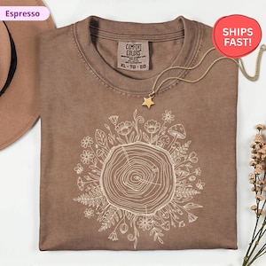 May include: Espresso-colored t-shirt with a white floral and tree ring design. The shirt has a crew neck and short sleeves. A gold star pendant necklace is draped over the shirt. The shirt is labeled "Comfort Colors" and is size XL-TG-EG.